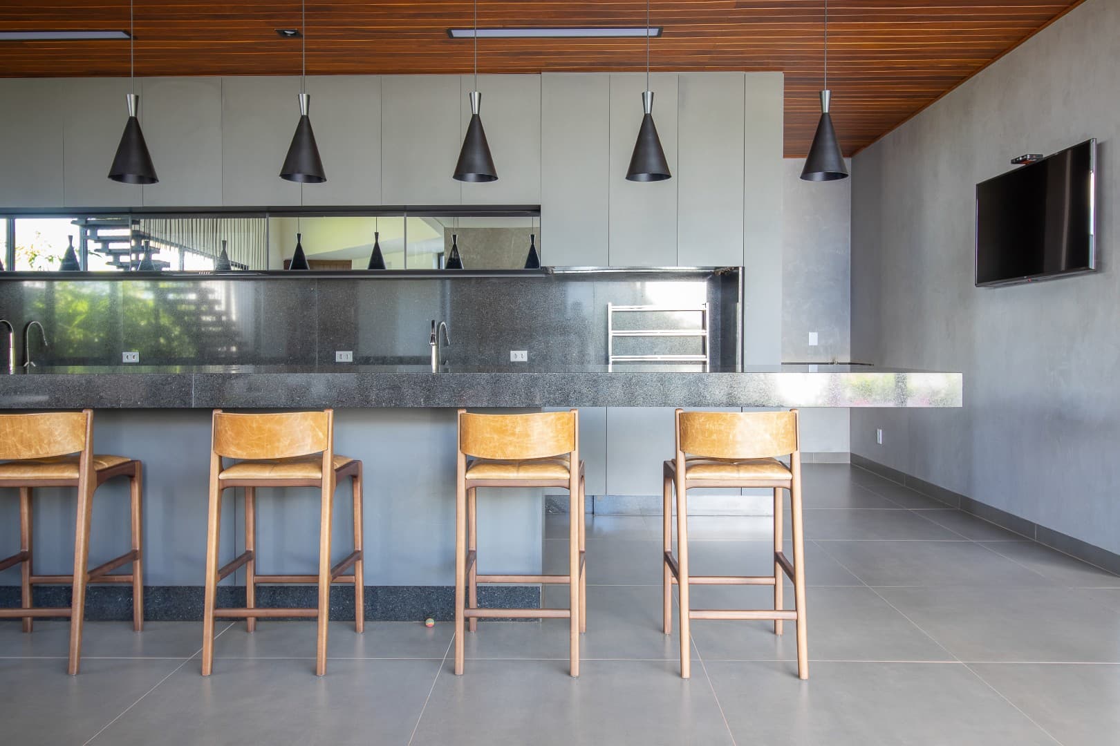 Casa CaCle – área gourmet com bancada em concreto, banquetas em madeira e TV